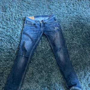Blåa jeans från Dondup - Snygga blå jeans från Dondup med slitningar och hål på låret för en cool, avslappnad look. Klassisk med kontrastsömmar och läderpatch bak i midjan. Jeansen har smal passform och är tillverkade i mjukt denimtyg. Byxorna är i ett bra skick tyvärr har själva lapen där det står George/ slingor lossnat som ni kan se i bild 5. Hör av er vid frågor Med vänlig hälsning, Anton