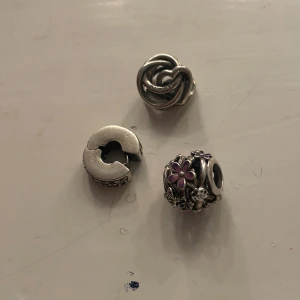  Pandora berlocker - Tre snygga Pandora berlocker i silver. En med lila blommor och fjärilar, en med hjärtformad knut där det står 'Always', och en glittrig klämberlock. Perfekt för att pimpa ditt armband med personliga detaljer och lite extra bling. Köpta förra året men har inte kommit till användning tyvärr.