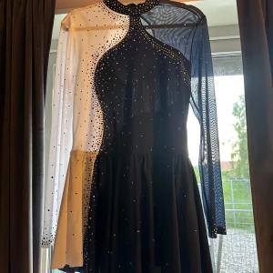 Mörkblå och vit glittrig konståkningsklänning  - Konståkningsklänning i svart och vitt med glittriga stenar och meshdetaljer. Klänningen har långärmade ärmar, en cool asymmetrisk design och hög hals med knappar. Använd en gång på träning då den inte passar mig i smaken och är för stor. Storlek L men skulle säga att den passar M då klänningen är liten i storlek. Pris kan diskuteras.