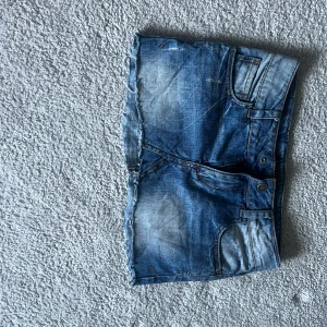 Blå jeanskjol från Sisley - Säljer en blå jeanskjol från Sisley med rå kant nertill och snygga bakfickor med broderade detaljer. Kjolen har klassisk femficksdesign och bälteshällor. Perfekt för dig som gillar en avslappnad och cool stil.