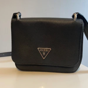 Svart axelväska från Guess i skinn - Snygg svart axelväska från Guess med klassisk trekantslogga i metall framtill. Väskan har justerbar axelrem och är tillverkad i slätt skinnmaterial. Perfekt storlek för det viktigaste och stilren design som passar till många outfits.