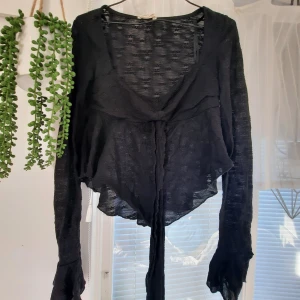 Svart omlottkofta med knyt - Svart tunn omlottkofta med långa ärmar och knyt framtill. Koftan har en lätt transparent look och asymmetrisk nederkant, perfekt för lager-på-lager och en edgy vibe.