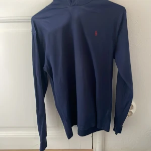 Marinblå hoodie Polo Ralph Lauren - Snygg marinblå hoodie från Polo Ralph Lauren med klassisk röd logga broderad på bröstet. Tröjan är i mjuk bomull och har en clean, stilren look som passar perfekt till jeans eller chinos.