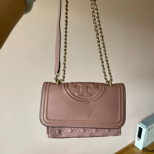 Rosa quiltad axelväska från Tory Burch - Supersnygg rosa axelväska från Tory Burch i skinn med quiltade detaljer och guldfärgad kedjerem. Väskan har magnetlås och präglad logga på locket. Perfekt storlek för det viktigaste och ger en lyxig touch till din outfit.