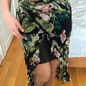 Blommig omlottkjol från Guess - Säljer en svart långkjol från Guess med tropiskt blommönster i grönt, rosa och vitt. Kjolen har omlottmodell och volangdetaljer längs nederkanten. Perfekt för dig som vill sticka ut med en färgstark och flowy look.