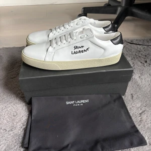 Vita Saint Laurent sneakers - Snygga vita sneakers från Saint Laurent med svart häl och broderad logga på sidan. Skorna har platt sula, rund tå och klassisk snörning. Tillverkade i lyxigt skinn och levereras med originalkartong och dustbag.