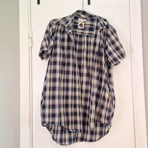 Rutig kortärmad skjorta från Kostym XS - Kortärmad skjorta från Kostym i rutigt mönster med blå, gul och beige toner. Lite längre bak och med fickor. Köpt själv från loppis och haft på bara några gånger, i väldigt bra skick. Storlek XS men jag tycker är ganska stor så borde passa till S med.