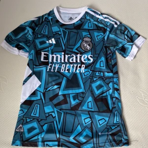 Real Madrid Vini Jr. Adidas fotbollströja - Snygg Real Madrid fotbollströja med VINI JR. och nummer 7 på ryggen. Tröjan har ett unikt blått och svart geometriskt mönster, vita detaljer och klubbmärke på bröstet. Tillverkad i lätt och ventilerande polyester med klassisk Adidas-logga och Aeroready-teknologi.