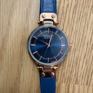 Blå damklocka med roséguldsdetaljer från KAT - Stilren damklocka med blått läderarmband och blå urtavla. Klockan har roséguldsfärgade detaljer och markerade index. Perfekt accessoar för att lyfta din outfit.
