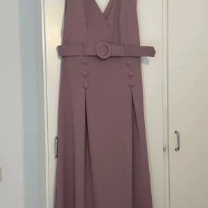 Ljuslila långklänning med bälte och knappar - Säljer en elegant ljuslila långklänning med djup v-ringning och bredt bälte i midjan. Klänningen har dekorativa knappar framtill och ett stilrent, ärmlöst snitt. Perfekt för speciella tillfällen!