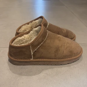 Bruna tofflor från 157 - Mysiga bruna tofflor från 157 med mjukt, fluffigt foder och ovandel i mocka. Perfekta att ha hemma under kyliga dagar. Sulan är platt och ger bra grepp på golvet. Går även att använda ute