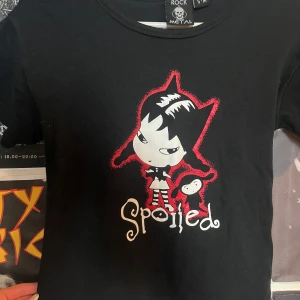Svart t-shirt med tryck från Rock Metal - Svart babytee med ett stort tryck av en tecknad figur med kattöron och texten 'Spoiled' i vitt och rött framtill. T-shirten har rund hals och korta ärmar. Perfekt för dig som gillar alternativ stil.
