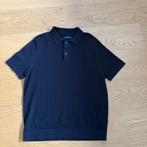 Svart pikétröja från Riley - Snygg svart pikétröja från Riley med klassisk krage och två knappar framtill. Tröjan har korta ärmar och är tillverkad i ett lätt, perforerat material som ger en sportig och fräsch känsla. Perfekt för dig som gillar stilrena och enkla plagg.