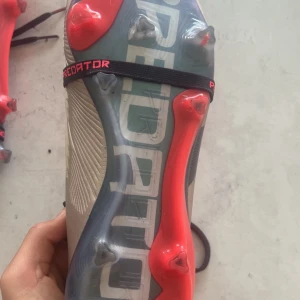 Adidas Predator fotbollsskor beige/röd - Snygga Adidas Predator fotbollsskor i beige med svarta detaljer och neonröda inslag på plös och dobbar. Skorna har snörning och är tillverkade i syntetmaterial för bästa grepp och känsla på planen. Perfekta för dig som vill sticka ut på matchen.