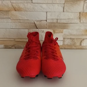 Nike Mercurial röda fotbollsskor - Snygga Nike Mercurial fotbollsskor i rött och orange med svart-vita detaljer. Skorna har snörning, högre skaft och mönstrad ovandel. Perfekta för dig som vill sticka ut på planen med stil och grym passform.