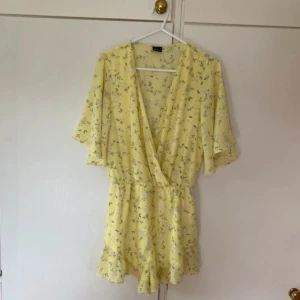 Gul blommig omlott playsuit med volang - Supersöt gul byxdress med blommigt mönster och omlott-ringning. Den har vida ärmar och volangdetaljer längst ner på shortsen samt resår i midjan för skön passform. Perfekt för sommaren!🌸🌸