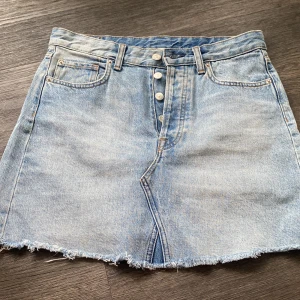 Ljusblå jeanskjol från H&M - Ljusblå jeanskjol från H&M i klassisk femficksmodell med knappar framtill och rå, fransig fåll. Kjolen har bälteshällor och bakfickor, perfekt för en avslappnad och trendig look.