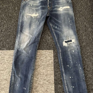 Dsquared2 blå slitna jeans, stl 44 - Snygga blå jeans från Dsquared2 i modell Skater Jean. Jeansen har slitna detaljer, hål och vit färgstänk för en cool, streetinspirerad look. Klassisk femficksmodell med smal passform och låg midja. Perfekta för dig som gillar edgy stil.