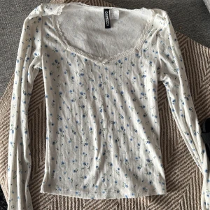 Blommig långärmad topp från H&M Divided - Supersöt långärmad topp från H&M Divided i vitt med små blå blommor. Toppen har ribbad struktur, spetskant vid halsringningen och en feminin v-ringad form. Perfekt för dig som gillar romantisk och söt stil.