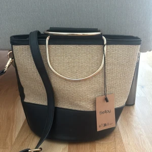 Beige och svart axelväska med metallhandtag - Snygg axelväska i beige och svart med stilrena metallhandtag och justerbar axelrem. Väskan har en modern design med struktur i materialet och detaljer i guld. Perfekt för att lyfta din outfit.