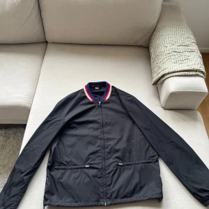 Svart jacka från Gucci - Säljer en svart jacka från Gucci med ett stort färgglatt tryck på ryggen. Jackan har långa ärmar och en ribbad krage med ränder i rött, vitt och blått. Perfekt för dig som vill ha något stilrent men ändå iögonfallande.