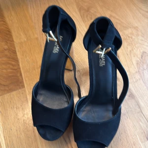 Svarta klackskor från Michael Kors - Säljer ett par svarta klackskor från Michael Kors med öppen tå och bred klack i guldig färg. Skorna har en elegant design med rem över foten och spänne i guld. Perfekta för dig som vill ha en stilren och lyxig look.