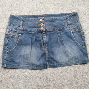 Kort Jeanskjol med knappar och fickor - Säljer en klassisk blå jeanskjol med tre dekorativa knappar framtill och både fram- och bakfickor. Kjolen har bälteshällor och är i en kort modell, perfekt till sommaren. Midjemått ca 46 cm och längd ca 29 cm.   Storlek XL men skulle säga L  . Säljs pågrund av att den är för liten. 