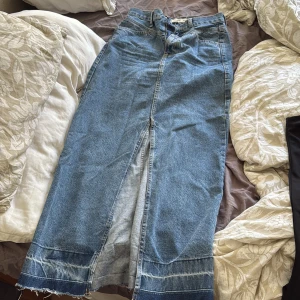 Lång jeanskjol med slits - Säljer en snygg lång jeanskjol i klassisk blå denim. Kjolen har en hög midja, markerade sömmar och en tydlig slits framtill för en cool look. Perfekt att styla med sneakers eller boots.