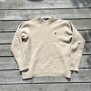 Beige ulltröja från Polo Ralph Lauren - Klassisk beige tröja från Polo Ralph Lauren i mjuk ull. Tröjan har rund halsringning, långa ärmar och en liten broderad logga på bröstet. Perfekt för lager-på-lager och enkel att matcha med det mesta.