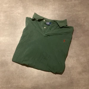 Grön pikétröja från Polo Ralph Lauren - Klassisk grön pikétröja från Polo Ralph Lauren med röd broderad logga på bröstet. Tröjan har korta ärmar, krage och knappar vid halsen. Perfekt för en avslappnad och stilren look. Tröjan är i storlek 2XB och är lite blekt. Tröjan är väldigt stor😁 det är bara att höra av sig vid frågor👍