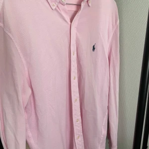 Ljusrosa skjorta från Ralph Lauren M - Säljer en snygg ljusrosa skjorta från Ralph Lauren i storlek M. Skjortan är i ett tunt mesh-liknande bomullstyg, har klassisk krage med knappar och den ikoniska broderade loggan på bröstet. Perfekt för dig som gillar stilrena och fräscha plagg.