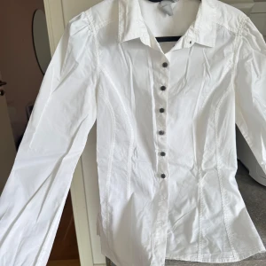 Vit långärmad skjorta med knappar - Klassisk vit skjorta med lång ärm och svarta knappar framtill. Skjortan har en figurnära passform och krage, perfekt för en clean och stilren look. 