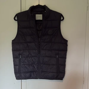 Svart dunväst från Jack & Jones - Snygg svart dunväst från Jack & Jones med quiltad design och hög krage. Västen har två dragkedjefickor framtill och stängs med dragkedja. Perfekt nu till sommaren och att slänga över en hoodie för en chill look. Västen är i väldigt bra skick och pris går att prata om. Västen är i storlek XS men jag skulle säga att den passar S bättre