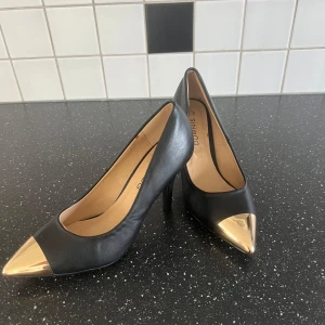 Svarta pumps med guldfärgad spets - Stilrena svarta pumps med hög klack och spetsig tå i guldfärgad metall-look. Skorna har en elegant form och är tillverkade i ett material som liknar skinn. Perfekta för att lyfta din outfit med en lyxig touch.