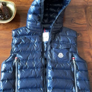 Mörkblå dunväst från Moncler - Snygg mörkblå dunväst från Moncler med glansig finish, huva och dragkedja framtill. Västen har två fickor med dragkedja och en bröstficka med lock och Moncler-logga. Perfekt för lager-på-lager och streetstyle.