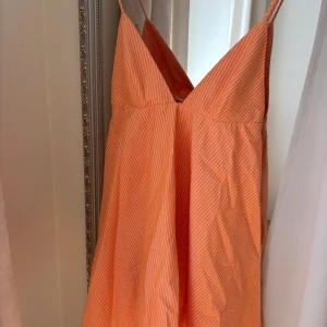 Orange randig långklänning med smala axelband - Säljer en orange långklänning med vita tunna ränder och smala axelband. Klänningen har v-ringad hals och är ärmlös, perfekt för varma dagar. Luftig och snygg modell som ger en somrig känsla.