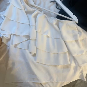 Vit offshoulder volangtop - Säljer en vit offshoulder topp med flera lager volanger. Toppen har en luftig och flowy känsla, perfekt för varma dagar. Snygg asymmetrisk axel och lätt material som ger en cool look.