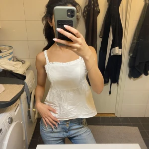 Vit volangig linnetopp med smock - Supersöt vit linnetopp med volangkant upptill och smockad rygg. Toppen har breda axelband och en lätt utsvängd passform som ger en luftig känsla. Perfekt till jeans eller kjol för en fräsch look.