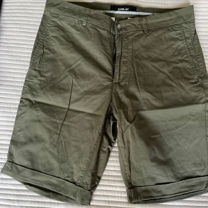 Olivgröna shorts från Replay - Snygga olivgröna shorts från Replay med klassisk passform och uppvikta benslut. Tillverkade i bomull med fickor fram och bak samt hällor för bälte. Perfekta för varma dagar och enkel att matcha med t-shirt eller skjorta.
