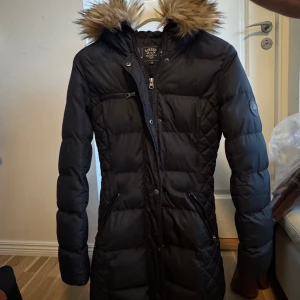 Svart lång dunjacka med päls - Lång svart dunjacka från Vinson Polo Club med snygg fuskpäls på huvan. Jackan har quiltad design, dragkedjor på fickorna och en extra bröstficka med dragkedja. Perfekt för kalla dagar med sin varma och stilrena look. Storlek 34-36.