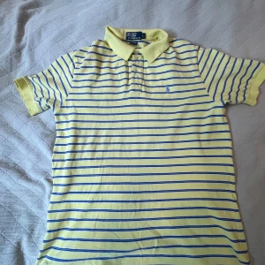 Gul och blå randig piké Polo Ralph Lauren - Snygg gul pikétröja från Polo Ralph Lauren med blå ränder och klassisk krage. Tröjan är i 100% bomull och har korta ärmar. Perfekt för dig som gillar preppy stil och vill sticka ut med färg i garderoben. Den har några fläckar men inget farligt.