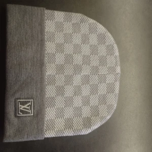 Grå rutig mössa med logotyp - Snygg grå mössa med rutigt mönster och uppvikt kant. På kanten finns en broderad logotyp. Perfekt för kyliga dagar och enkel att matcha med olika outfits.