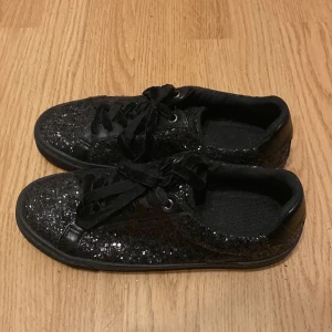 Svarta glittriga sneakers med snörning - Säljer ett par svarta sneakers med glittrig yta och svarta skosnören. Skorna har rund tå och platt sula, perfekta för dig som vill sticka ut lite extra. Passar till många olika outfits och ger en cool touch till din stil.