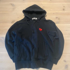 CDG ziphoodie - Svart hoodie med dragkedja och huva från Comme des Garçons Play. Röd hjärtlogga på bröstet och stort tryck på ryggen. Mjuk bomullskänsla och fickor framtill. Skicka ett meddelande för frågor, storlek, mått osv. 
