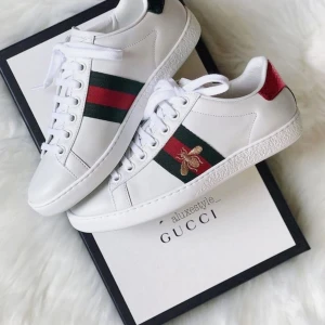 Vita sneakers från Gucci med broderad bi - Säljer ett par vita sneakers från Gucci med gröna och röda ränder på sidan samt en broderad guldfärgad bi. Skorna har vit snörning och detaljer i rött och grönt på hälen. Klassisk och stilren modell.