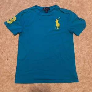 Blå t-shirt från Polo Ralph Lauren - Snygg blå t-shirt från Polo Ralph Lauren med stort gult broderat logotyptryck på bröstet och siffran 3 på ärmen. Klassisk rund hals och korta ärmar. Perfekt för en avslappnad stil. Denna Polo Ralph Lauren tröja har lite större märke än de vanliga Polo tröjorna. Skriv till mig om ni har nån fråga eller nått om tröjan tröjan  eller om storlek för jag vet inte exakt vad storleken på tröjan är!