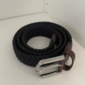 Marinblått flätat bälte från John Henric - Snyggt marinblått flätat bälte med bruna skinn-detaljer och silverfärgat spänne från John Henric. Perfekt accessoar för att lyfta din outfit med en stilren touch. Helt oanvänt! Nypris:500kr| 100cm långt, bara höra av sig vid frågor!