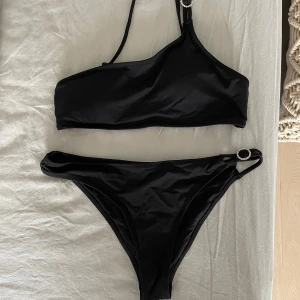 Svart bikini från Zara med asymmetrisk topp - Snygg svart bikini från Zara med en axelbandstopp och dekorativa ringdetaljer i silver på både topp och underdel. Topp med justerbara band och klassisk bikiniunderdel. Använd fåtal gånger vid solning men ej bad. (det går att köpa bara underdelen eller överdelen om det önskas) 😊
