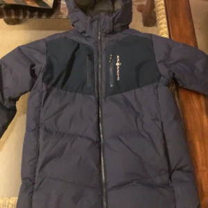 Mörkblå dunjacka från Sail Racing - Säljer en mörkblå dunjacka från Sail Racing med huva och dragkedja. Jackan har en bröstficka med dragkedja och en patch på ärmen med texten 'Sail Antarctica Expedition'. Perfekt för kalla dagar.