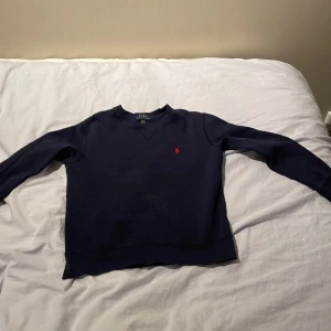 Mörkblå sweatshirt från Polo Ralph Lauren - Mörkblå sweatshirt från Polo Ralph Lauren med klassisk rund halsringning och röd broderad logga på bröstet. Tröjan har långa ärmar och ribbade muddar vid ärmslut och nederkant. Perfekt för en avslappnad stil.